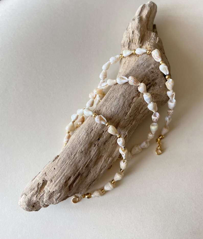 May include: A delicate necklace made of small, cream-colored seashells and gold-colored beads. The necklace is displayed on a piece of driftwood, showcasing its natural and beachy aesthetic. The necklace has a gold clasp.
