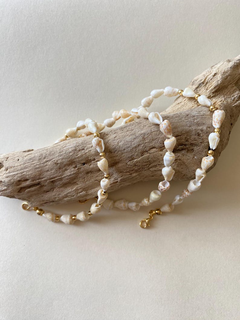 May include: A delicate necklace made of small, cream-colored seashells and interspersed with gold-colored beads. The necklace is displayed on a piece of driftwood against a neutral background. The necklace has a gold-colored clasp.