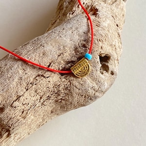 Puede incluir: Un collar de cordón rojo con un pequeño colgante dorado en forma de semicírculo con un patrón de líneas concéntricas texturizadas y una cuenta única, redonda y de color turquesa brillante, situada junto al colgante dorado.