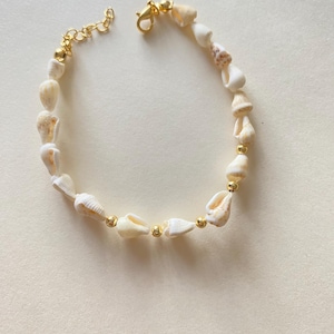 May include: A delicate seashell bracelet featuring small, cream-colored shells strung together with gold-colored beads and a gold-tone chain clasp. The bracelet is designed for a comfortable fit and adds a touch of coastal charm to any outfit.