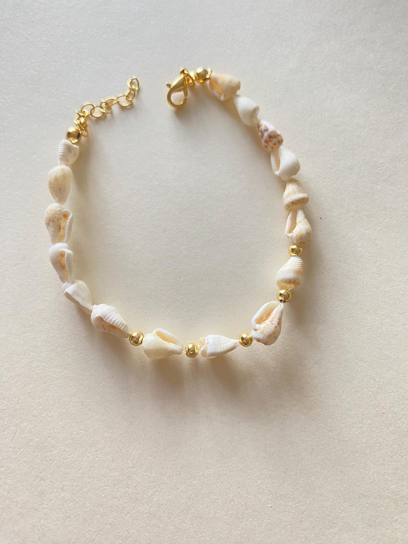 May include: A delicate seashell bracelet featuring small, cream-colored shells strung together with gold-colored beads and a gold-tone chain clasp. The bracelet is designed for a comfortable fit and adds a touch of coastal charm to any outfit.
