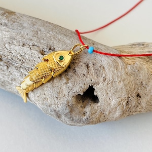 Op de afbeelding: Een goudkleurige vishangerketting met een rood koord. De vishanger heeft een textuuroppervlak en een groene edelsteen oog. Een kleine blauwe kraal accentueert het rode koord.