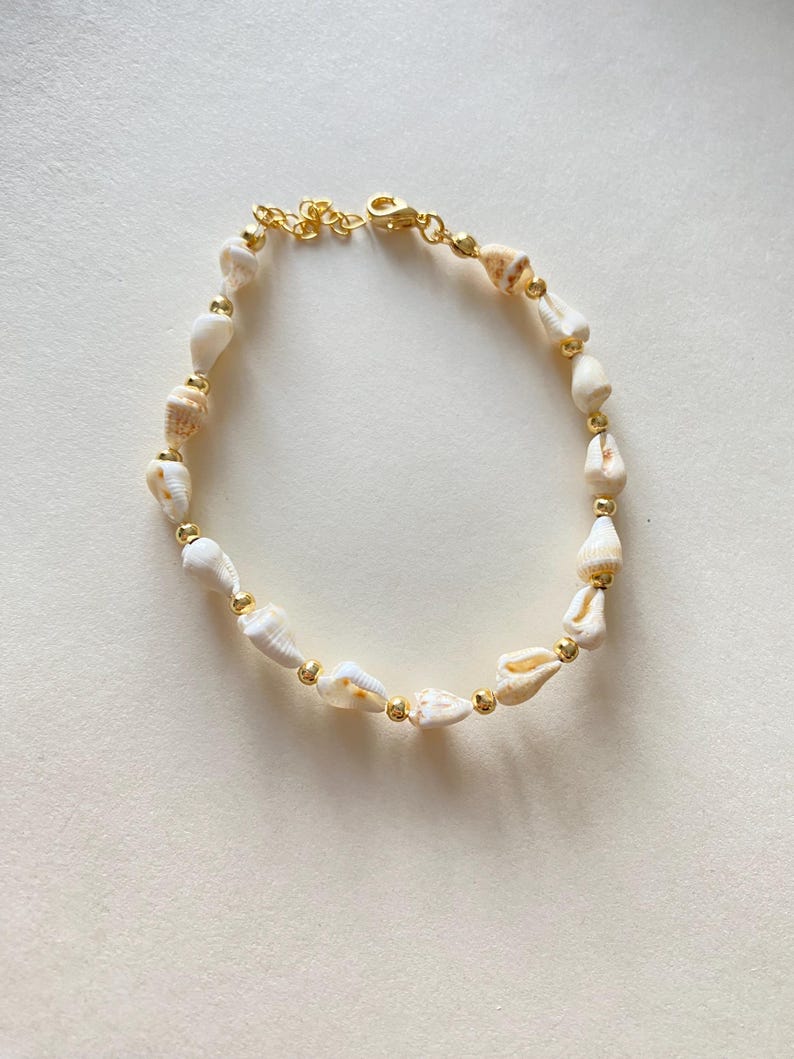 May include: An anklet featuring natural seashells and small gold-colored beads strung on a delicate gold-tone chain. The shells are primarily white with tan markings. The anklet has a lobster clasp closure.