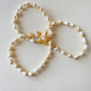 May include: A necklace and two bracelets made of small, white seashells and gold-colored beads. The jewelry has gold-colored clasps and chain extenders. The seashells have natural tan and brown markings. The jewelry is displayed on a light beige surface.
