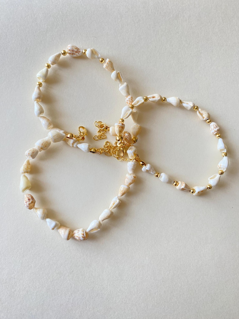 May include: A necklace and two bracelets made of small, white seashells and gold-colored beads. The jewelry has gold-colored clasps and chain extenders. The seashells have natural tan and brown markings. The jewelry is displayed on a light beige surface.