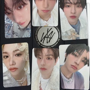 May include: A collection of six Stray Kids photo cards featuring close-up portraits of the band members. The cards are arranged on a black surface, with the Stray Kids logo visible on one card.