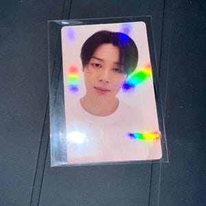 Può includere: Una fototessera olografica in una custodia di plastica trasparente. La tessera mostra una persona con i capelli scuri e una maglietta bianca. La luce arcobaleno si rifrange sulla superficie della tessera.