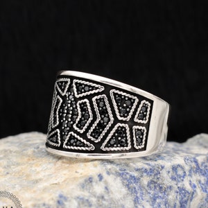 May include: A silver ring with a geometric design. The ring features a black inlay with a pattern of triangles and other shapes, filled with small black stones. The ring has a polished finish and a wide band.
