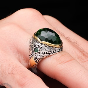 Może przedstawiać: Srebrny pierścionek z dużym zielonym kamieniem szlachetnym i złotymi akcentami. Pierścionek ma szczegółowy wzór z skrzydłami i mniejszym zielonym kamieniem szlachetnym.