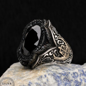 May include: A silver ring featuring a large black gemstone surrounded by smaller black gemstones. The ring has intricate silver and gold detailing, including a design of scales and a bird.