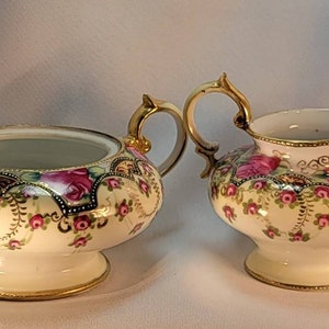 May include: A vintage porcelain sugar bowl and creamer set with a floral design. The set features a white background with pink and green flowers and gold accents.