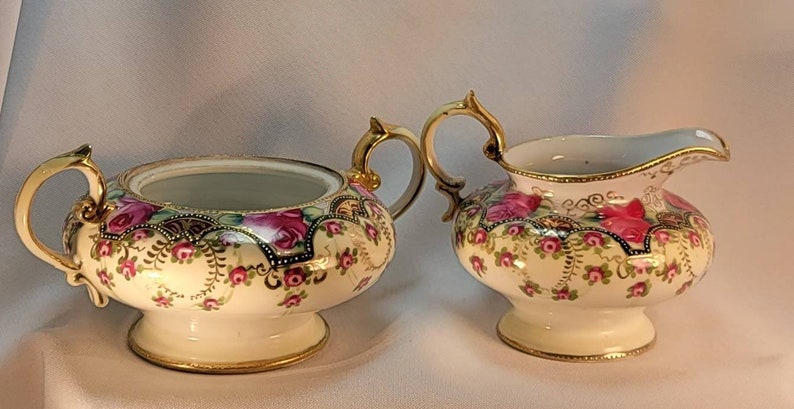 May include: A vintage porcelain sugar bowl and creamer set with a floral design. The set features a white background with pink and green flowers and gold accents.