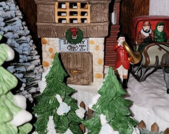 May include: A miniature village scene with a green-roofed building with a sign that reads "Sammy Bean & Son". There are two small evergreen trees with snow on them and a miniature figure playing a horn.