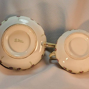 May include: Bottom view of a white porcelain creamer and sugar bowl set with a floral design. The bottom of the creamer has a gold rim and the text "Zsolnay" in gold lettering.