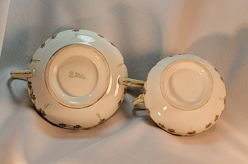 May include: Bottom view of a white porcelain creamer and sugar bowl set with a floral design. The bottom of the creamer has a gold rim and the text "Zsolnay" in gold lettering.