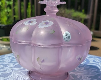 May include: A pink glass candy dish with a scalloped edge and a floral design. The dish is hand-painted with purple flowers and green leaves. The dish has a lid with a knob on top. The dish is made by Fenton and says "Handpainted in USA" on the bottom.