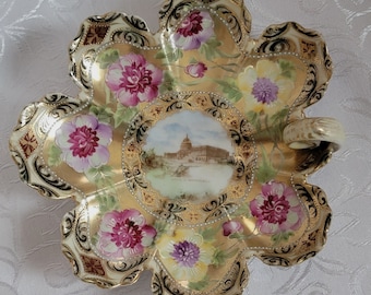 May include: A decorative, flower-shaped dish with a gold rim and floral design. The dish features hand-painted pink and yellow flowers, with a central image of a building. The dish has a handle and is likely made of porcelain.