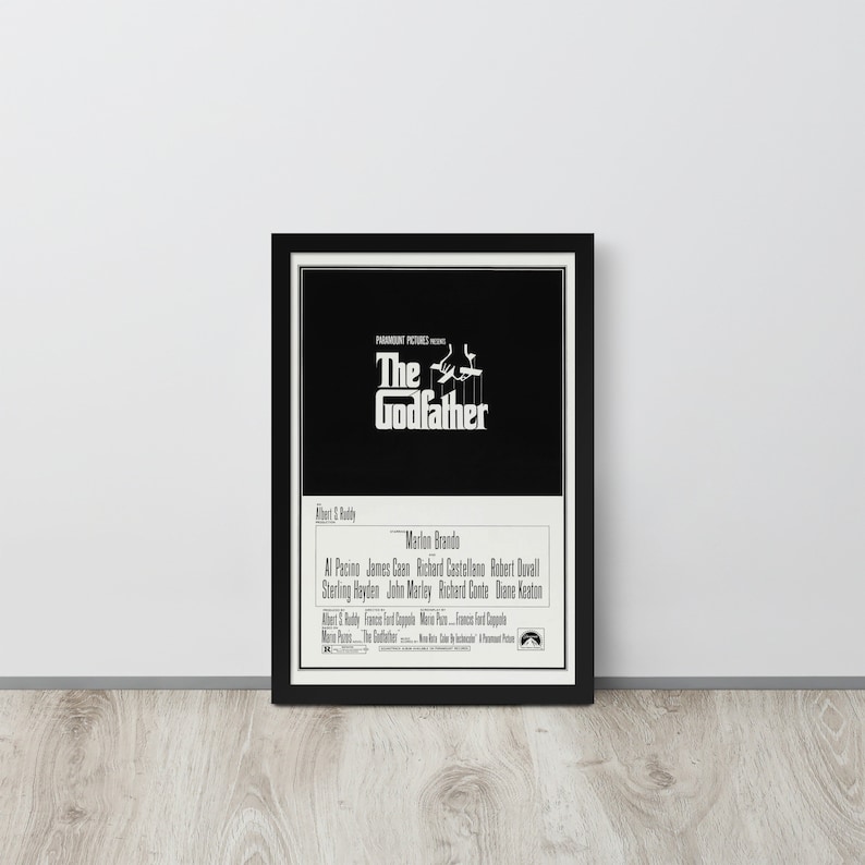 May include: A black and white movie poster for the film "The Godfather" featuring the title in bold white text and a silhouette of a man holding a puppet. The poster also includes the names of the actors, including Marlon Brando, Al Pacino, and James Caan.