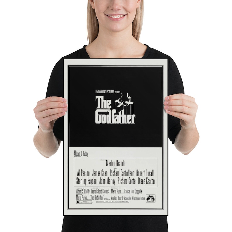 May include: A black and white poster for the movie "The Godfather" featuring the film's title and a silhouette of a hand holding strings with puppets. The poster also includes the names of the actors and director.
