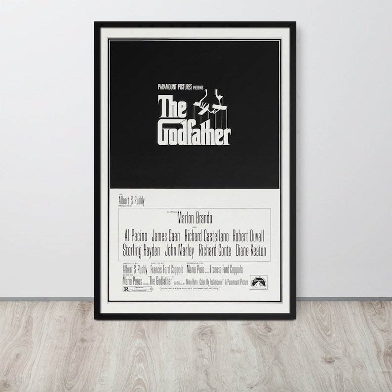May include: Black and white movie poster for The Godfather, featuring a hand holding strings with puppets. The text "The Godfather" is in a large, bold font. The actors' names are listed below, including Marlon Brando, Al Pacino, and James Caan.