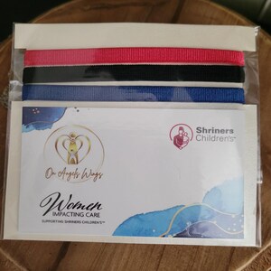 May include: A set of three ribbon bracelets in red, black, and blue. The bracelets are packaged in a clear plastic bag with a card that reads "On Angels Wings Women Impacting Care Supporting Shriners Children's".