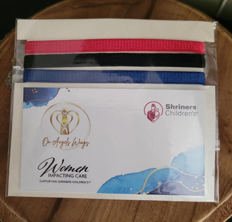 May include: A set of three ribbon bracelets in red, black, and blue. The bracelets are packaged in a clear plastic bag with a card that reads "On Angels Wings Women Impacting Care Supporting Shriners Children's".