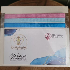May include: A white card with a blue and white watercolor background. The card features a logo with the text "On Angels Wings" and "Women Impacting Care Supporting Shriners Children's". The card also features a logo for Shriners Children's with the text "Shriners Children's". The card is packaged with three ribbons: pink, light blue, and blue.