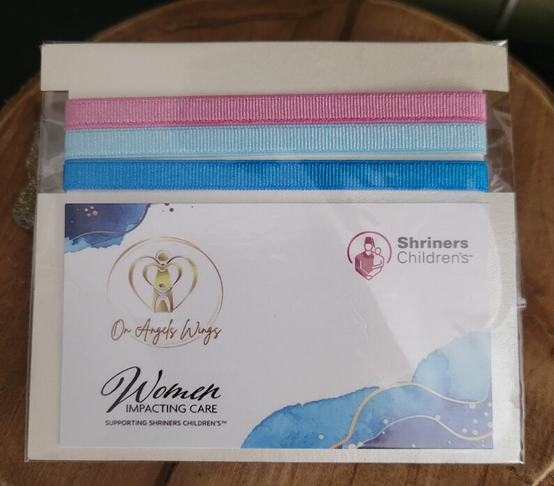 May include: A white card with a blue and white watercolor background. The card features a logo with the text "On Angels Wings" and "Women Impacting Care Supporting Shriners Children's". The card also features a logo for Shriners Children's with the text "Shriners Children's". The card is packaged with three ribbons: pink, light blue, and blue.