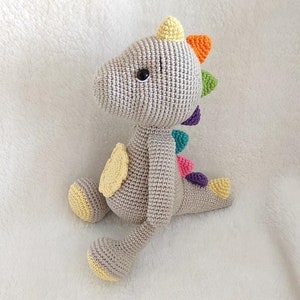 May include: A gray crocheted dinosaur toy with yellow, orange, green, pink, and purple accents on its back. The dinosaur has a yellow crocheted horn on its head and black eyes.