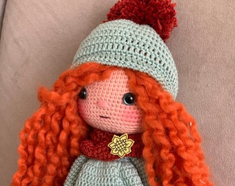 May include: A crocheted doll with long, curly red hair, wearing a light blue dress, a red scarf, and a light blue hat with a red pom-pom. The doll has a flower-shaped brooch on its chest.