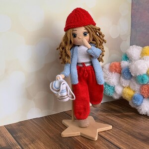 May include: A crocheted doll with long brown hair, wearing a red hat, a blue cardigan, a white shirt, and red pants. The doll is holding a small blue and white crocheted bag.