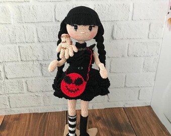 May include: A crocheted doll with black hair, a black dress, and a red bag with a jack-o'-lantern face. The doll is wearing black and white striped socks and black shoes.