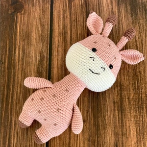 May include: A pink crocheted giraffe toy with brown ears and a white face. The giraffe has black eyes and a stitched smile.