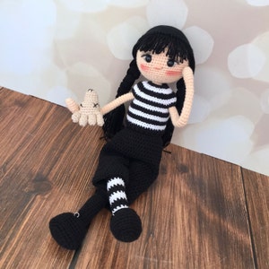 K&ouml;nnte beinhalten: Eine geh&auml;kelte Puppe mit schwarzen Haaren, einem schwarz-wei&szlig; gestreiften Shirt, einem schwarzen Rock und schwarz-wei&szlig; gestreiften Str&uuml;mpfen. Die Puppe h&auml;lt ein kleines geh&auml;keltes Spielzeug.