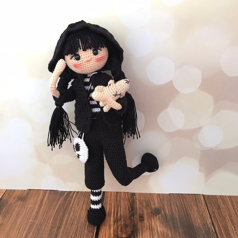 K&ouml;nnte beinhalten: Eine geh&auml;kelte Puppe mit schwarzen Haaren, die ein schwarz-wei&szlig; gestreiftes Shirt, eine schwarze Hose und schwarz-wei&szlig; gestreifte Socken tr&auml;gt. Die Puppe h&auml;lt ein geh&auml;keltes wei&szlig;es Tier.