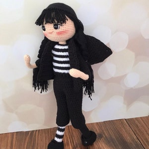 K&ouml;nnte beinhalten: Eine geh&auml;kelte Puppe mit schwarzen Haaren, einem schwarz-wei&szlig; gestreiften Shirt, schwarzen Hosen und wei&szlig;-schwarz gestreiften Socken. Die Puppe tr&auml;gt schwarze Schuhe und hat eine schwarze geh&auml;kelte Jacke.