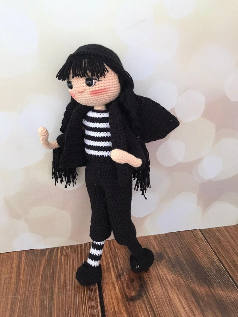 K&ouml;nnte beinhalten: Eine geh&auml;kelte Puppe mit schwarzen Haaren, einem schwarz-wei&szlig; gestreiften Shirt, schwarzen Hosen und wei&szlig;-schwarz gestreiften Socken. Die Puppe tr&auml;gt schwarze Schuhe und hat eine schwarze geh&auml;kelte Jacke.