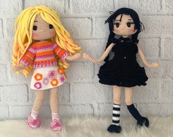 May include: Two crocheted dolls, one with blonde hair wearing a pink and orange striped sweater and a white skirt with pink flowers, and the other with black hair wearing a black dress with a ruffled hem and black and white striped stockings.