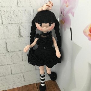 May include: A crocheted doll with black hair, a black dress, and black and white striped socks. The doll is wearing a small crocheted animal on its head.