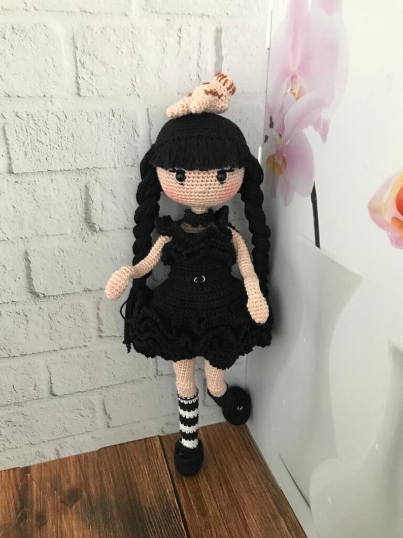 May include: A crocheted doll with black hair, a black dress, and black and white striped socks. The doll is wearing a small crocheted animal on its head.