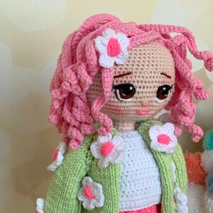 May include: A crocheted doll with pink curly hair, a green cardigan with white flowers, and a white shirt. The doll has brown eyes and pink lips.