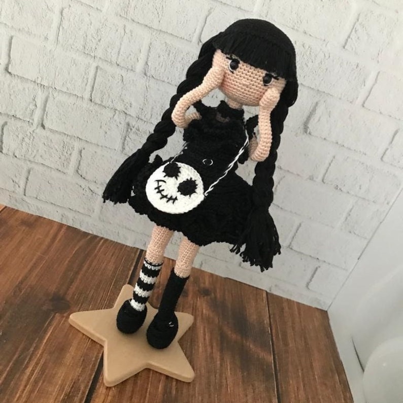 May include: A black crocheted doll with long black braids, wearing a black dress with a white skull-shaped purse. The doll is standing on a wooden star-shaped base.