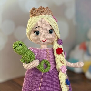 May include: A handmade crochet doll of a princess with long blonde hair, a crown, and a purple dress. The doll is holding a green chameleon and a green ring. The hair is decorated with colorful flowers.