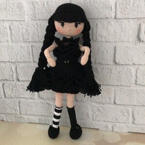 May include: A crocheted doll with black hair, a black dress, and black and white striped socks. The doll is standing on a white brick background.