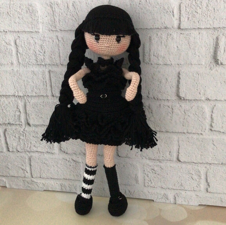 May include: A crocheted doll with black hair, a black dress, and black and white striped socks. The doll is standing on a white brick background.