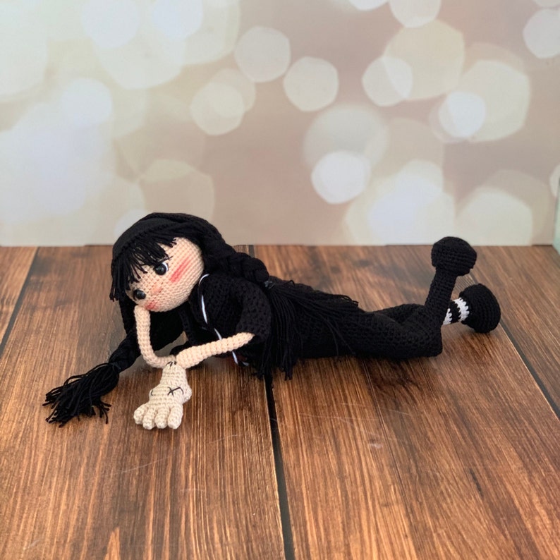 K&ouml;nnte beinhalten: Eine schwarze geh&auml;kelte Puppe mit langen schwarzen Haaren, die ein schwarzes Outfit mit wei&szlig;en Akzenten tr&auml;gt, liegt auf einer Holzoberfl&auml;che.
