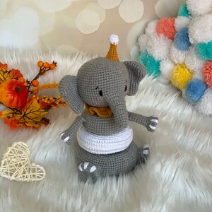 May include: A handmade grey crochet elephant toy with a yellow collar and party hat. The elephant has white accents and is sitting on a white surface. Decorative autumn foliage and a heart-shaped decoration are in the background.
