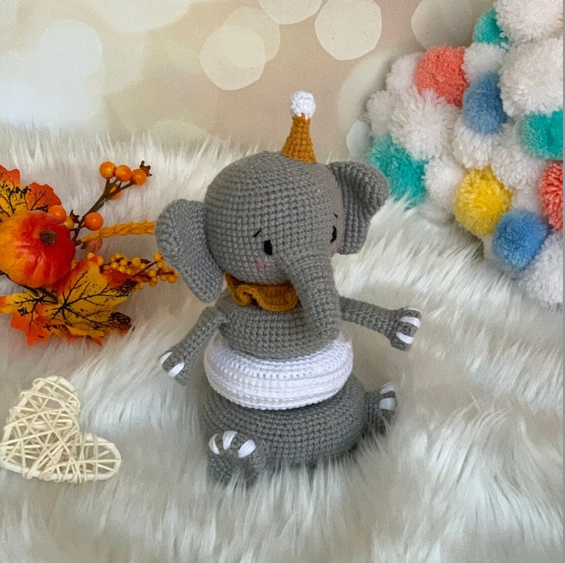 May include: A handmade grey crochet elephant toy with a yellow collar and party hat. The elephant has white accents and is sitting on a white surface. Decorative autumn foliage and a heart-shaped decoration are in the background.