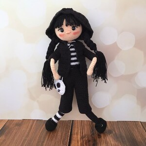 K&ouml;nnte beinhalten: Eine schwarze H&auml;kelpuppe mit einem schwarz-wei&szlig; gestreiften Shirt, schwarzen Hosen und schwarz-wei&szlig; gestreiften Socken. Die Puppe hat schwarze Haare, eine schwarze Kapuzenjacke und ein wei&szlig;es Sch&auml;del-Accessoire.