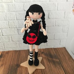 May include: A crocheted doll wearing a black dress with a red pumpkin-shaped purse. The doll has black hair in pigtails and is holding a crocheted hand.